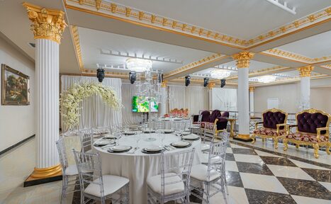Арендовать Банкетный зал “Император”. Банкетный зал с колоннами, декорированными золотом. Округлый стол с прозрачными стульями, сервировка столовых приборов. Люстра на потолке, черно-белая плитка на полу, обитые бархатом кресла. Настенные картины, светлые шторы и декор цветами.