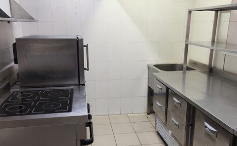 Арендовать Buon Gusto (Хороший вкус). Кухонное пространство с плиткой на полу и стенах. Металлическое оборудование: духовка, плита с четырьмя конфорками, рабочие столы с ящиками и раковиной. Две полки над столешницей. Светлое освещение.