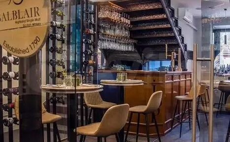 Арендовать Лофт с кухней площадью 80 м². Барное пространство с деревянной стойкой, винными стеллажами под лестницей. Круглые столы с высокими стульями. Стеклянные перегородки, подвесные бокалы. Нейтральные и тёплые цвета. Уютное освещение. Текст на круглой табличке.