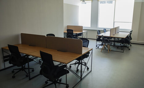 Арендовать Smart Coworking(Умный Коворкинг). Просторный офис с двумя группами столов. Каждая группа включает два стола с перегородками и черными офисными креслами. Светлые стены, большие окна, обеспечивающие естественное освещение. Пол светлый, потолок белый.