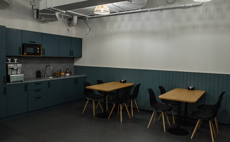 Арендовать Smart Coworking(Умный Коворкинг). Небольшая кухня с синими шкафами и серой столешницей. Имеется микроволновая печь, кофемашина и раковина. Две деревянные столы с черными стульями. Белые стены с синим панелированием. Потолок с системой вентиляции и подвесными светильниками.