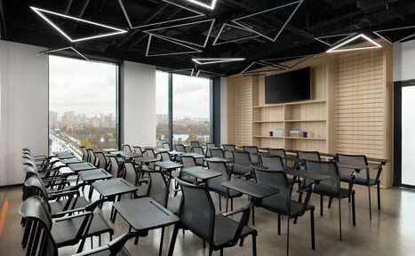 Арендовать YES Technopark(ДА Технопарк). Конференц-зал. Современное офисное пространство с большим количеством стульев и столиков для конференций. Черные потолочные светильники геометрической формы, светлые деревянные панели на стене с экраном, большие окна с видом на город.