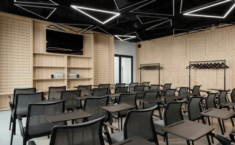 Арендовать YES Technopark(ДА Технопарк). Конференц-зал. Конференц-зал с рядами черных сетчатых кресел и столиков. Светлая деревянная отделка стен, TV-экран, полки с водой, открытая вешалка. Геометрические светильники на черном потолке, нейтральная цветовая гамма.