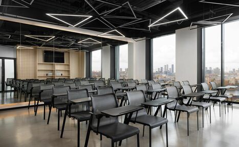 Арендовать YES Technopark(ДА Технопарк). Конференц-зал. Аудиторийное помещение с рядами серых стульев с откидными столиками, большими окнами с видом на город, зеркальной стеной и деревянной отделкой. Потолочные светильники геометрической формы. Пол бетонный, цветовая гамма нейтральная.