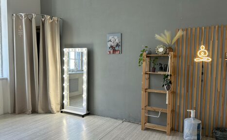 Арендовать Лофт СВЕТОСФЕРА. Просторная комната с серыми стенами, зеркалом с подсветкой, шторами, деревянной полкой с растениями и декором, неоновым светильником в форме медитирующей фигуры, ламинированным полом, окном слева, картина на стене.