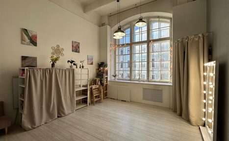Арендовать Лофт СВЕТОСФЕРА. Светлое помещение с высоким потолком, большая арочная стеклянная дверь, подвесные светильники. На стенах картины, полка с вазами и растениями, складные стулья. Справа зеркало с лампочками и шторы. Светлые деревянные полы.