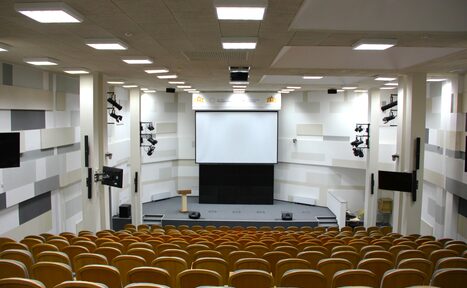 Арендовать Технопарк «Университетский». Аудиторное пространство со ступенчатыми рядами деревянных кресел, сцена с трибуной и большим экраном, два проектора по бокам, многоуровневое потолочное освещение, звуковая аппаратура по стенам, бело-серые стены с акустическими панелями.