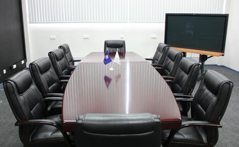 Арендовать Конференц-зал площадью 17000 м² до 280 гостей. Большой конференц-зал с деревянным столом и десятью черными кожаными креслами. На столе маленькие флаги. На полу темный ковролин. На заднем плане большой экран на подставке. Вертикальные жалюзи на окне. Белые стены.