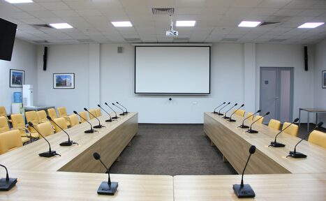 Арендовать Технопарк «Университетский». Зал «Эволюция». Конференц-зал с U-образной расстановкой столов, желтыми креслами, микрофонами, проектором и экраном. Белые стены, подвесной потолок с потолочными панелями и встроенными светильниками. В углу кулер для воды. На стенах картины.