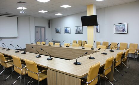 Арендовать Технопарк «Университетский». Зал «Эволюция». Конференц-зал с двумя рядами деревянных столов и микрофонами, окружённый жёлтыми стульями. На задней стене висят картины. Потолок с встраиваемыми светильниками. На стене установлен телевизор. Слева проекционный экран. Тёмно-серый ковролин.