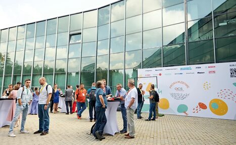 Арендовать Технопарк «Университетский». Открытая площадка.. Открытое пространство с вымощенной плиткой, вдоль здания с высокими стеклянными окнами. Люди стоят у высоких столов с белыми скатертями. На заднем плане белый баннер с логотипами и разноцветными графическими элементами.