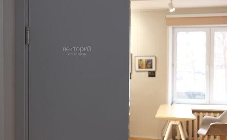 Арендовать Лекторий Научно-просветительского центра Музея М.А. Булгакова. Лекторий с деревянным столом и стулом у окна. Белые стены, на них картины. Деревянный пол. Естественное и точечное освещение. На двери надпись: "лекторий lecture room". Светло-бежевые и белые цвета.