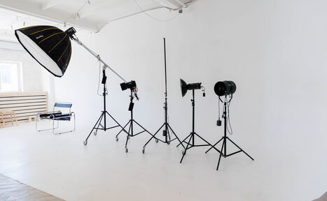 Арендовать Art Room Studio (Арт Рум). Фотостудия. Студийное пространство для фотосъёмки, белые стены и пол, профессиональное осветительное оборудование на стойках, светодиодные лампы, оконный проем с естественным светом, небольшая скамья, стул с металлическим каркасом, светлая древесина на полу.