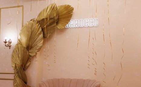 Арендовать Барокко. Элегантный лофт. Золотые декоративные листья на стене, неоновая надпись, розовый диван с закругленными формами, светло-персиковая стена. Вертикальные золотые ленты спускаются с потолка. Настенный светильник в классическом стиле.