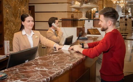Арендовать Амбассадор Отель. ```plaintext
Стойка регистрации с мраморной поверхностью, сотрудница в бежевом костюме вручает документ мужчине в красном свитере. На заднем плане другой сотрудник использует телефон. В помещении люстра и декор на стене.
```