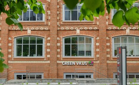 Арендовать Green Loft(Зеленый Лофт). Красное кирпичное здание с большими арочными окнами. Вывеска со словами "GREEN VKUS CAFE" на фасаде. Перед зданием металлографитовый забор и каменные ступени. Деревья на переднем плане.