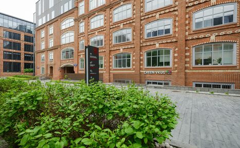Арендовать Green Loft(Зеленый Лофт). Красное кирпичное здание с большими арочными окнами. На переднем плане тротуар из серых плит и кусты. Видно черную стойку с текстом. Вывеска "GREEN VKUS" на фасаде здания. Обстановка ухожена, пешеходная зона.