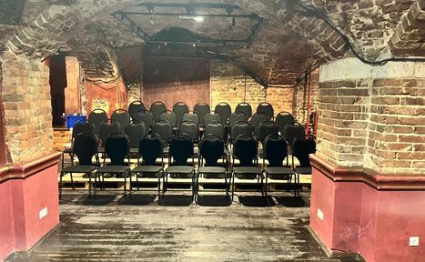 Арендовать Культурный центр «Хитровка». "Арт-зал". Старинное подвальное пространство с кирпичными арками и каменными стенами, темный деревянный пол, черные стулья в ряды, мягкое освещение сверху, бордовые панели на стенах, черные трековые светильники.