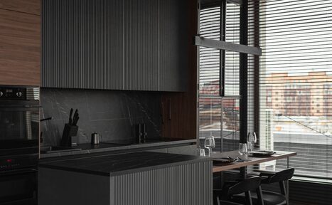 Арендовать Зал Black. Фотостудия Мимика. Современная кухня с серыми и черными элементами, барной стойкой и столом у окна. На столе три стеклянных бокала и приборы. Вертикальные жалюзи на окнах. Тёмные шкафы с деревянными вставками. Слева у стены плита и раковина.