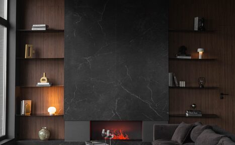 Арендовать Зал Black. Фотостудия Мимика. Гостиная с камином. По бокам камина расположены полки с книгами и декоративными элементами. На полу стоит стеклянный столик с бокалами. Справа тёмный диван. Стены облицованы тёмным деревом и чёрным мрамором. Освещение создают настольные лампы и окно слева.
