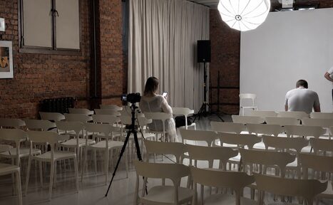 Арендовать Ö Art Space(Арт Спейс). Современное арт-лофт пространство. Лофт-пространство с кирпичными стенами, белыми стульями, расположенными рядами. Освещение от крупного подвесного светильника. На стене картинная рамка. Белая занавеска, светлая напольная поверхность. На штативе установлен фотоаппарат.