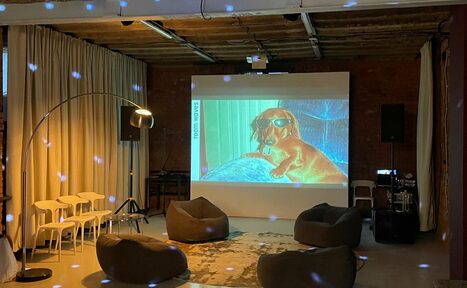 Арендовать Ö Art Space(Арт Спейс). Современное арт-лофт пространство. Лофт-пространство с кирпичными стенами и открытым потолком. В центре проектор с изображением собаки в очках. Мягкие кресла, белые стулья, торшер, светодиодное освещение. Шторы и ковёр светлых тонов. Звуковое оборудование, стол, провода.