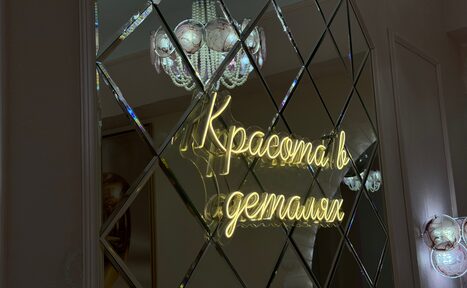 Арендовать Лофт «Голд Эпл». Зеркальная стена с ромбовидными панелями, неоновая надпись "Красота в деталях", люстра с круглыми элементами, светлые стены.