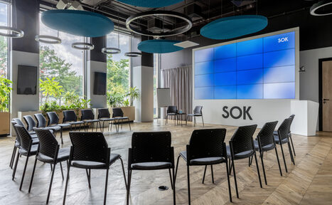 Арендовать SOK Рыбаков Тауэр. Просторная конференц-зал с большим экраном на стене, перед которым расположены стулья полукругом. Пол деревянный, потолок с круглыми акустическими панелями. Стены с окнами и зелеными растениями. Надпись «SOK» на стойке под экраном.