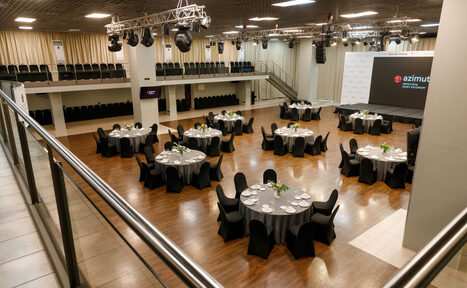 Арендовать Зал «Азимут». Большое многоуровневое конференц-пространство с паркетным полом. Круглые столы с темными скатертями расставлены по залу, окруженные стульями. Есть сцена, экран и система освещения на потолке. Стены светлые, балкон с сиденьями.