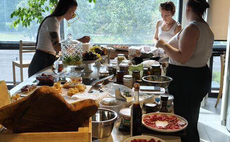 Арендовать Пространство СемЬСюР с Кухней. Просторная комната с панорамным окном, стол с разнообразными продуктами и посудой. Несколько человек стоят вокруг стола. Светлый интерьер, много естественного света. Видно зелень за окном, через стекло. Деревянные и стеклянные элементы.
