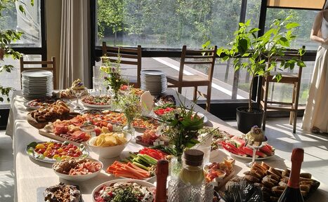Арендовать Пространство СемЬСюР с Кухней. Просторная комната с большими окнами и видом на зелень. Белый стол с разнообразными блюдами, включая овощи и закуски. На столе стеклянные графины и бутылки. Четыре деревянных стула, растения в горшках. Естественное освещение.