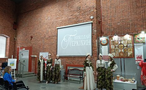 Арендовать Выставочный зал Форум. Пространство с кирпичными стенами, серым полом, сценической зоной. На сцене группа людей в костюмах. Проектор, экран с текстом. По периметру стулья для зрителей. Выставочные витрины с экспонатами. Светлое освещение.