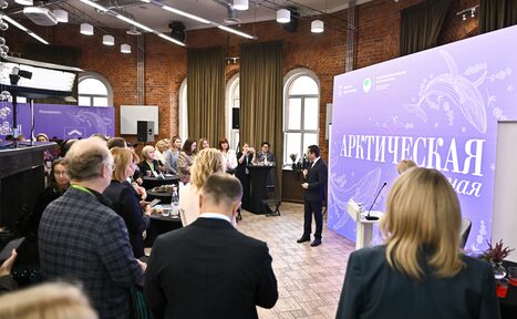 Арендовать Зал на 9-м этаже Водонапорной башни. Просторное помещение с кирпичными стенами и высокими арочными окнами с коричневыми шторами. Помост с фиолетовым баннером, микрофон на стойке. Люди стоят, слушают выступающего. Точечное освещение, пол покрыт плиткой.