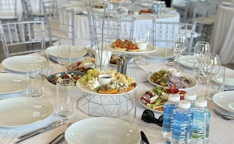 Арендовать Event  Hall ARCOBALENO (Эвент Холл АРКОБАЛЕНО) . Столовая зона с круглым столом, покрытым белой скатертью, сервированная тарелками, приборами и стеклянной посудой. На столе закуски и бутылки воды. Прозрачные стулья, светлый интерьер, круглые столы на заднем плане.