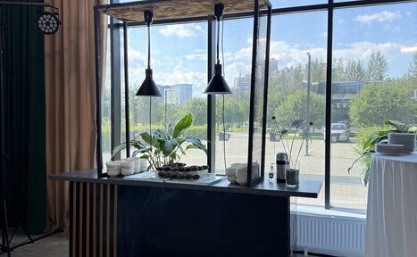 Арендовать Event  Hall ARCOBALENO (Эвент Холл АРКОБАЛЕНО) . Барная стойка у окна, черные подвесные светильники, темно-синяя глянцевая поверхность с деревянными элементами, белые тарелки и чашки, стеклянная ваза с растениями, стол на белой скатерти, вид на улицу через большое окно.