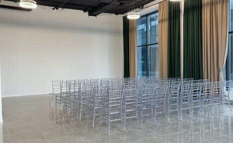 Арендовать Event  Hall ARCOBALENO (Эвент Холл АРКОБАЛЕНО) . Просторное помещение с белыми стенами и большими окнами, закрытыми зелеными и бежевыми шторами. Расставлены прозрачные пластиковые стулья в рядах. Потолочные светильники обеспечивают освещение. Пол выложен светлой плиткой.