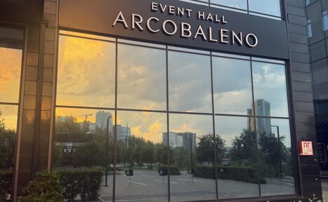 Арендовать Event  Hall ARCOBALENO (Эвент Холл АРКОБАЛЕНО) . Фасад здания с большими зеркальными окнами, отражающими городские пейзажи и закатное небо. Чёрная табличка с текстом "EVENT HALL ARCOBALENO". Состояние фасада ухоженное, внизу зелёные кусты.