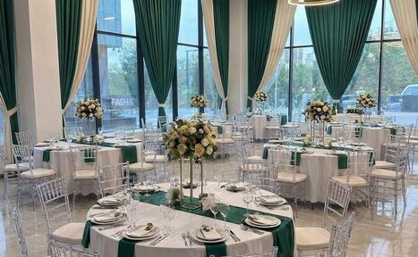 Арендовать Event  Hall ARCOBALENO (Эвент Холл АРКОБАЛЕНО) . Просторный зал с круглыми столами, покрытыми белыми скатертями и зелеными дорожками. Прозрачные стулья вокруг столов. На столах сервировка и цветочные композиции. Высокие окна с зелеными и белыми шторами. Светлые стены и пол, подвесной светильник.
