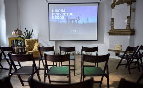 Арендовать Артиста Лекторий. Пространство с деревянным полом, белыми стенами и экраном для проектора. Ряды черных складных стульев, на экране текст. У стены кресло, зелёное растение и светильник. Справа здание с декоративной плиткой.