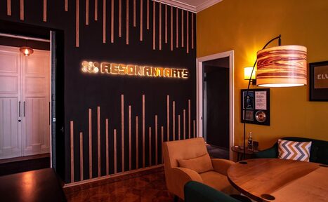 Арендовать Resonant Arts – студия звукозаписи и видео съёмки. Современное пространство с черной стеной и деревянными вставками, надписью "Resonamate". Оранжевое кресло и деревянный стол. Желто-оранжевая стена с постером и квадратным светильником. Паркетный пол и круглые светильники.