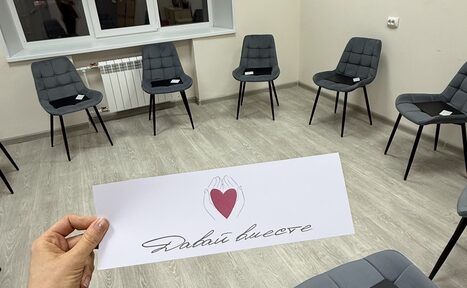 Арендовать Давай Вместе. Комната с круговой расстановкой серых стульев на светлом деревянном полу. У стены белая батарея под подоконником. В руке держат бумагу с красным сердцем и текстом. Светлые стены и спокойное освещение.