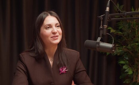 Арендовать Подкастная А40. Студийное пространство с затемненной драпировкой, крупная микрофонная установка, на заднем плане растение. Женщина в темном пиджаке с розовой брошью сидит перед микрофоном. Освещение мягкое.