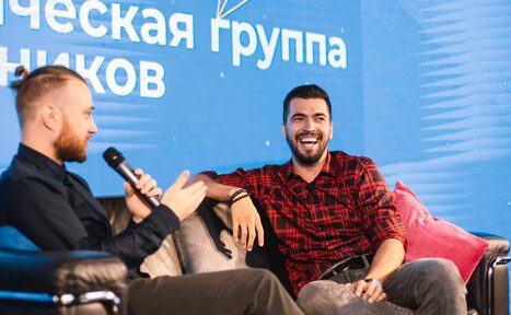 Арендовать Медиапространство «Маросейка 3/13». Гостиная зона с двумя мужчинами на кожаном диване. Мужчина справа в красно-черной рубашке, мужчина слева с микрофоном. На фоне синий экран с текстом на русском. Подушки на диване.