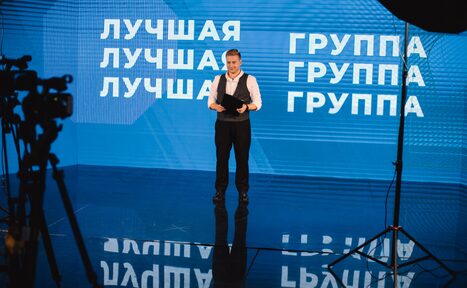 Арендовать Медиапространство «Маросейка 3/13». Студийное пространство с синим фоном и надписями "ЛУЧШАЯ ГРУППА". На переднем плане человек с бумагами, освещение из софитов, камера на штативе. Пол глянцевый.