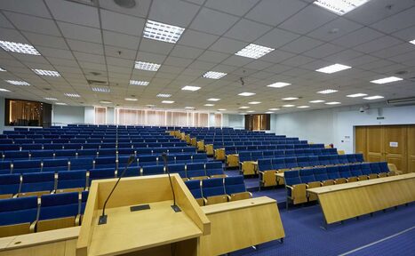 Арендовать Конференц-зал Петровский. Аудитория с рядами синих кресел и деревянными столами, кафедра спереди. Потолочные светильники равномерно освещают помещение. Стены светлого оттенка, дверь справа. Пол синие, коврового типа покрытия.
