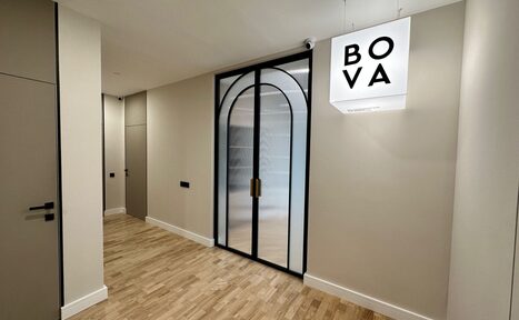 Арендовать Лекторий на 53 этаже Москва-Сити - 100 м², до 35 человек. Коридор с деревянным полом и светлыми стенами, стеклянная дверь с арочной конструкцией и черной рамой, рядом светящийся белый куб с черной надписью "BOVA". На дверях черные ручки. Освещение встроенное.