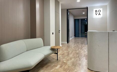 Арендовать Лекторий на 53 этаже Москва-Сити - 100 м², до 35 человек. Современное офисное пространство с паркетным полом, светлыми стенами и потолком. Слева мягкий диван с небольшим встроенным столиком, справа бетонная стойка. В глубине виден стеклянный вход. Освещение точечное. На стене знак с текстом "BOVA".