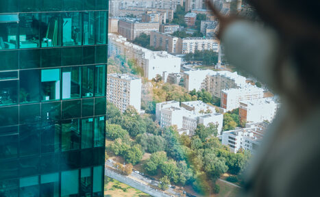 Арендовать Лекторий на 53 этаже Москва-Сити - 100 м², до 35 человек. Вид из окна высотного здания с отражением офиса на стекле. Внутри видны подсвеченные окна с офисной мебелью. За окном городской пейзаж с многоэтажными домами и деревьями. На переднем плане размытый силуэт человека.