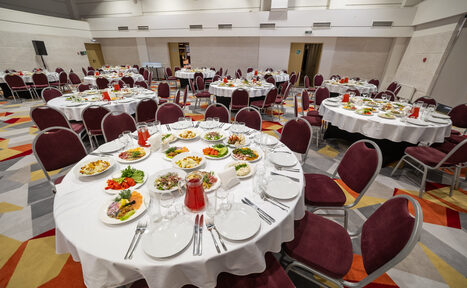 Арендовать Gorskiy City Hotel. Большой банкетный зал с круглой расстановкой столов, накрытых белыми скатертями. На столах находятся блюда с закусками, столовые приборы и стаканы. Стулья с бордовыми сиденьями, ковровое покрытие с геометрическим узором.