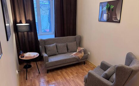 Арендовать Кабинет площадью 15 м² для работы. Небольшая комната с диваном и креслом серого цвета, столиком и торшером. Стены светлые, на одной картинка с растением. Окно закрыто тёмными шторами. Пол паркетный. На столике блокнот и ручки.